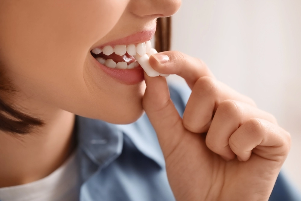 Close-up of a smiling person placing a white chewing gum piece into their mouth. Bright teeth and relaxed expression suggest freshness and satisfaction.